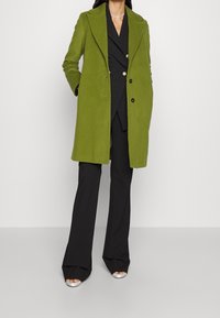 Grüner Mantel aus Wollmischung mit Reverskragen, doppelreihiger Vorderseite und Seitentaschen, kombiniert mit einem schwarzen, maßgeschneiderten Anzug und silbernen Absätzen.