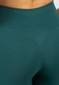 Zielone legginsy w wysokim stanie, wykonane z gładkiego, elastycznego materiału, z prążkowanym pasem i detalem w kształcie litery V.