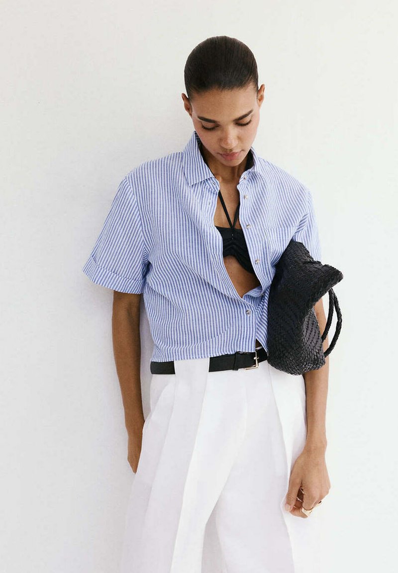 Femme portant une chemise à manches courtes rayée bleue par-dessus un haut noir, pantalon blanc avec une ceinture noire, tenant un sac tissé noir.
