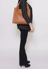 Borsa tote in pelle marrone con doppi manici, superficie texturizzata e forma rettangolare. Indossata insieme a pantaloni neri aderenti e top a fantasia.