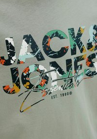 Sivi majica sa više bojama, isprskanom tipografijom s natpisima "JACK & JONES Essentials" i "EST. 1990", naglašavajući odvažne dizajnerske elemente.