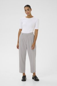 Femme debout en haut à manches courtes blanc, pantalon taille haute gris clair et mocassins noirs sur un fond clair uni.