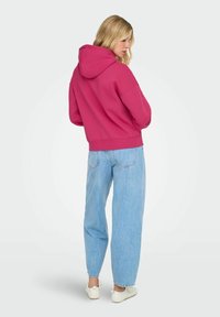 Une femme aux cheveux blonds porte un sweat à capuche rose, un jean bleu clair et des baskets blanches, se tenant dos à un fond gris uni.