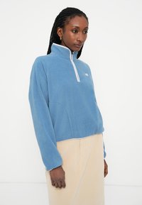 Pulover din fleece de culoare albastru deschis, cu guler înalt și fermoar până la jumătate, având o margine albă și detalii cu logo pe piept. Textură moale și croiala lejeră.