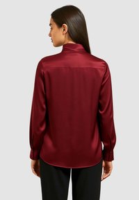 Camicia a maniche lunghe in satin bordeaux con colletto alto, texture liscia e vestibilità su misura; presenta un orlo leggermente curva e polsini con bottoni.