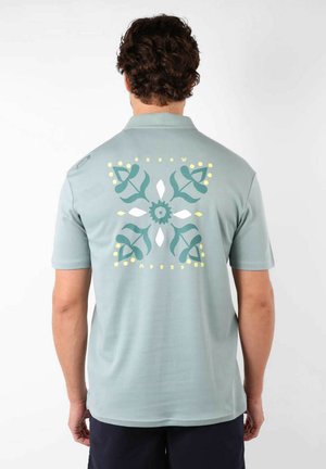 Polo shirt bleu clair à manches courtes avec un grand motif floral au dos en vert et jaune, avec une texture douce et lisse.