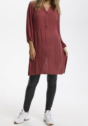 Vínové tunikové šaty s knoflíkovou légou a dlouhými rukávy, doplněné lesklými černými legínami a bílými sportovními botami. Hladká texturu látky.