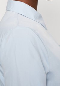 Camicia azzurro chiaro con colletto, caratterizzata da una texture liscia e un design aderente. Il tessuto della camicia sembra leggero con sottili dettagli di cucitura.