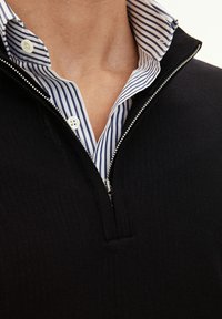 Svart zip-neck tröja med en texturerad stickad design, lagd över en blå och vit randig skjorta med krage. Vita knappar synliga.