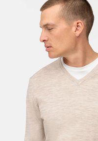 Maglione leggero beige a V con una texture morbida, caratterizzato da sottili variazioni di tonalità, indossato sopra una maglietta bianca a girocollo.