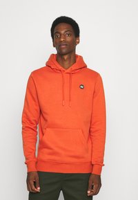 Mężczyzna w jaskrawopomarańczowej bluzie z kapturem z kieszenią z przodu i małym czarnym logo, stojący na tle jednolitego jasnego tła.