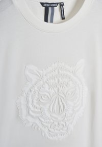 Camiseta blanca de corte regular con diseño de cara de tigre en relieve en el frontal; etiqueta de ropa negra con el nombre de la marca y la talla en el interior del cuello.