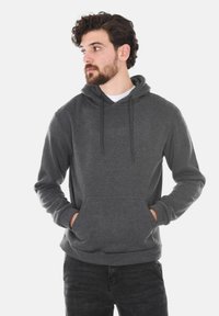 Felpa grigia realizzata in tessuto morbido, con tasca a marsupio, cappuccio regolabile con cordini e polsini a coste. Indossata sopra una maglietta bianca.