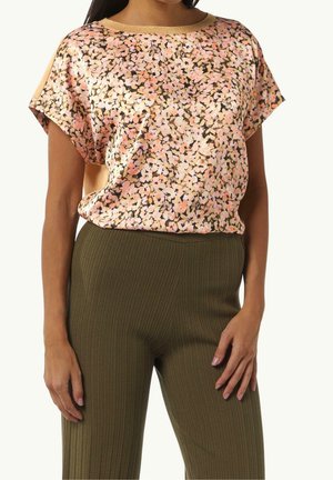 Vrouw die een kortemouwen blouse draagt met een bloemmotief in roze, perzik en zwart, gecombineerd met hooggetailleerde olijfgroene geribde broek.