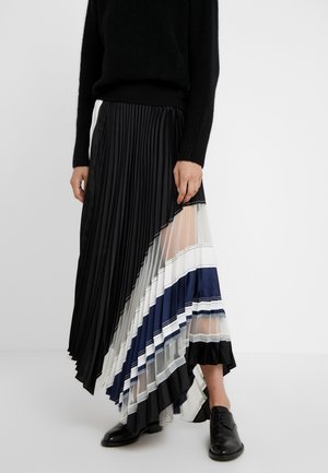 Persona que lleva un suéter negro y una falda plisada asimétrica con paneles en negro, blanco, azul marino y transparente, combinada con zapatos oxford negros.