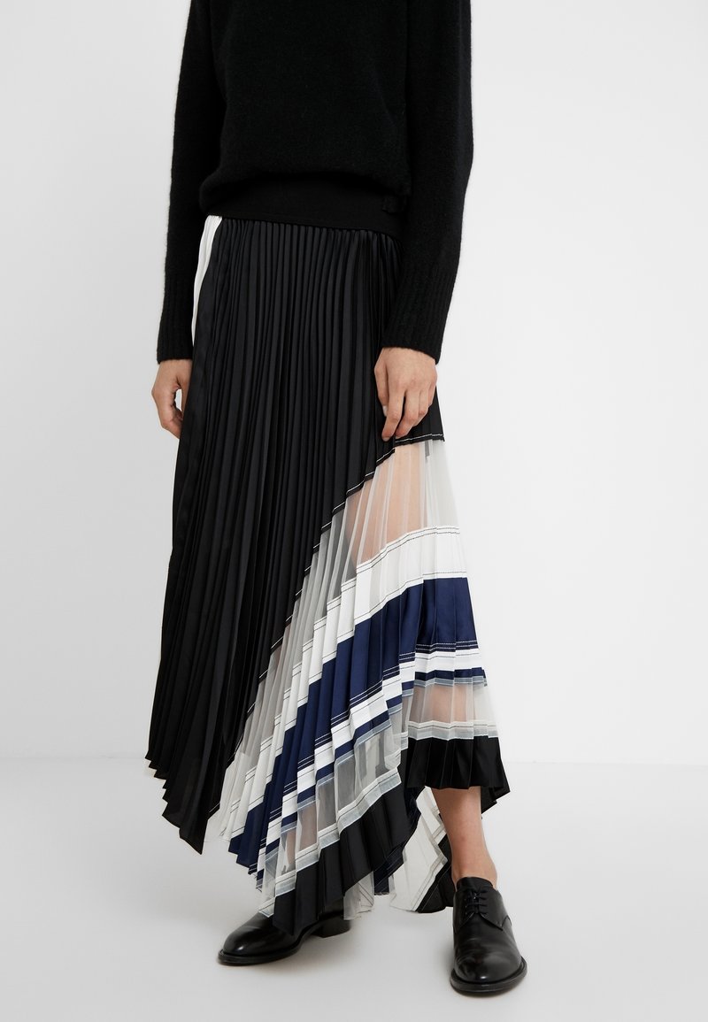 Personne portant un pull noir et une jupe plissée asymétrique avec des panneaux noirs, blancs, bleu marine et transparents, associée à des chaussures oxford noires.