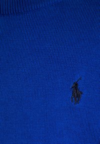 Modrý pletený svetr s vroubkovanou texturou, na levé straně zdobený malým černým vyšitým logem koně a jezdce.