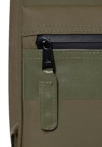 Olivgrüner Stoffbeutelabschnitt mit einem schwarzen Reißverschluss, strukturiertem Material, kontrastierenden Nähten und einem befestigten Lederzuglasche.