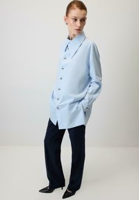 Gaiši zila pārliela krekls ar lielu apkakli un metāla pogām, kombinēts ar tumši zilām biksēm ar plašām kājsaitēm un melniem smailiem apaviem.