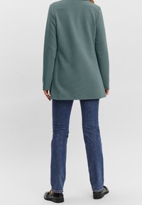 Personne portant une veste verte à manches longues, un jean bleu et des mocassins noirs, debout, dos tourné vers le spectateur sur un fond uni.