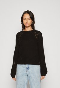 Zwarte gebreide trui met een geribbelde ronde halslijn, lange mouwen en opengewerkte details op de schouders. Gepaard met lichtblauwe jeans.