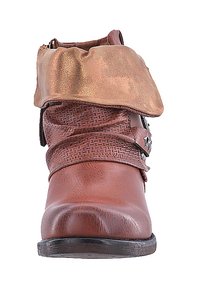 Bota de tobillo de cuero marrón con una correa trenzada texturizada y un puño metálico dorado. Presenta una puntera redondeada y una suela baja negra.