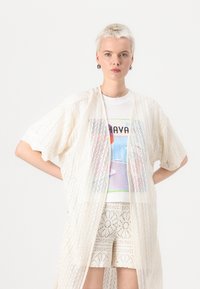 Kimono din dantelă în alb ofop, cu un model texturat în diamant, purtat peste un tricou grafic multicolor și pantaloni scurți cu model.