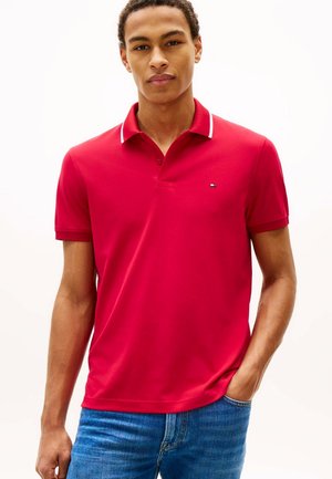 Giovane uomo che indossa una polo rossa con bordini bianchi e jeans blu, in piedi con una mano in tasca su uno sfondo uniforme.