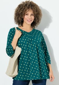 Cyan tuniek met een ontspannen pasvorm, 3/4 mouwen en kleine gele bloemenpatronen. Gecombineerd met een lichtbeige handtas.