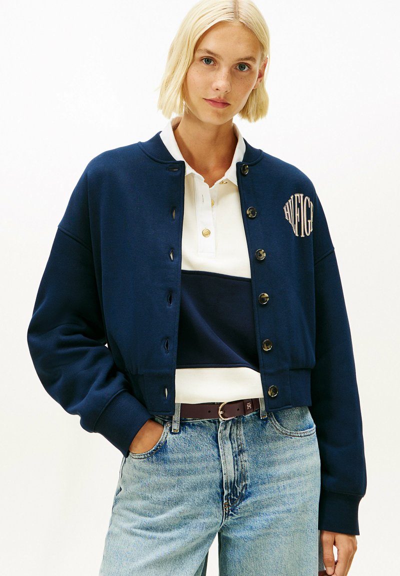 Mujer rubia que lleva una chaqueta azul marino abotonada sobre una camisa polo blanca y azul marino, pantalones vaqueros azul claro y un cinturón oscuro, de pie con una mano en el bolsillo.