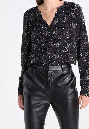Kobieta w czarnej bluzce z długim rękawem w kwiatowy wzór, wpuszczonej w wysoki stan czarnych skórzanych spodni, stoi na jednolitym tle.