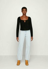 Top preto de mangas longas com um detalhe em forma de buraco, combinado com jeans retos de tom azul claro e botas de tacão em tom de pele, contra um fundo neutro.