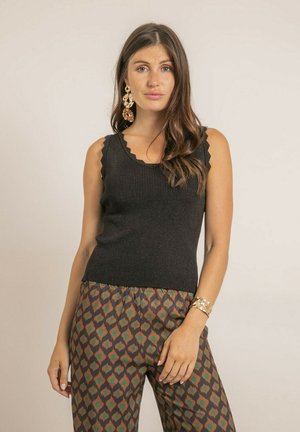 Femme aux longs cheveux bruns portant un haut noir sans manches, un pantalon à motifs marron et bleu-vert, de grandes boucles d'oreilles dorées et un bracelet en or.