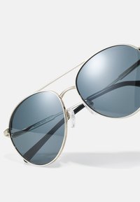 Lunettes de soleil aviateur avec monture en métal argenté, verres teintés bleus et embouts de branches noirs. Elles sont équipées de plaquettes de nez en silicone transparent pour un confort optimal.