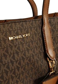 Borsa marrone con texture e logo dorato "MICHAEL KORS", con manici in pelle liscia e una chiusura metallica. I motivi creano un'eleganza sottile.