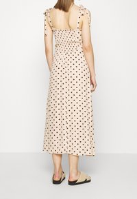 Vestido maxi de tela beige con lunares marrones, espalda fruncida y tirantes atados. Presenta un corte relajado y una silueta fluida.