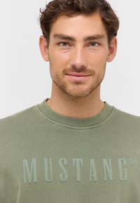 Sweat-shirt vert olive avec un col ras du cou côtelé et une marque texturée "MUSTANG" dans une couleur contrastante sur le devant.