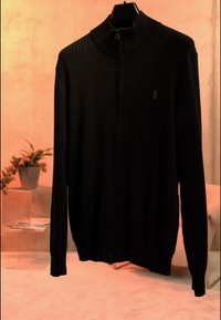 Pull noir côtelé à fermeture éclair avec col montant suspendu à un cintre noir, fond beige avec une plante dans un pot gris sur une table d'appoint transparente.