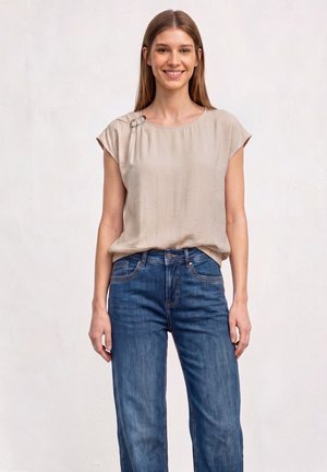 Jeune femme aux longs cheveux bruns portant un haut beige à manches courtes et un jean bleu, souriante et debout devant un fond blanc uni.