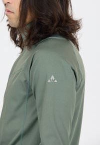 Mann mit langen dunklen Haaren, der eine grüne Jacke mit Reißverschluss trägt, auf dem oberen Arm befindet sich ein kleines weißes dreieckiges Logo.