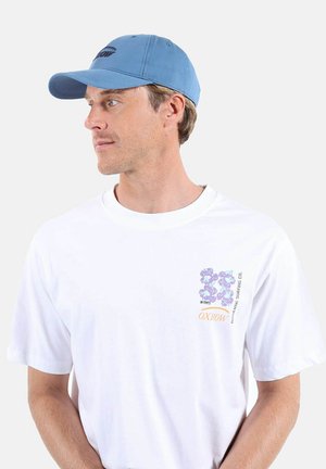 Homme portant une casquette bleu clair et un T-shirt blanc avec un motif floral violet et le texte orange "OXBOW" sur la poitrine, regardant vers la gauche.