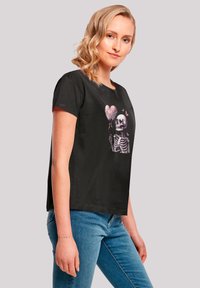 Black t-shirt with a graphic design featuring a skeleton holding a pink heart balloon, short sleeves, and a relaxed fit.