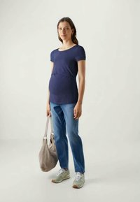Blusa de maternidade de manga curta em azul-marinho com detalhe franzido na cintura, combinada com jeans azul claro e ténis cinzentos, segurando uma mala bege.