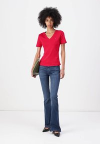 Raudonas V formos iškirptės marškinėliai su trumpomis rankovėmis, derinami su mėlynomis platėjančiomis džinsomis ir juodais batais, laiko žalią rankinę. Lygus audinio raštas.