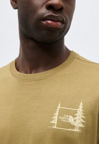 Primer plano de una persona que lleva una camiseta de color mostaza con un logotipo blanco de "The North Face" y un gráfico de árbol en el pecho.