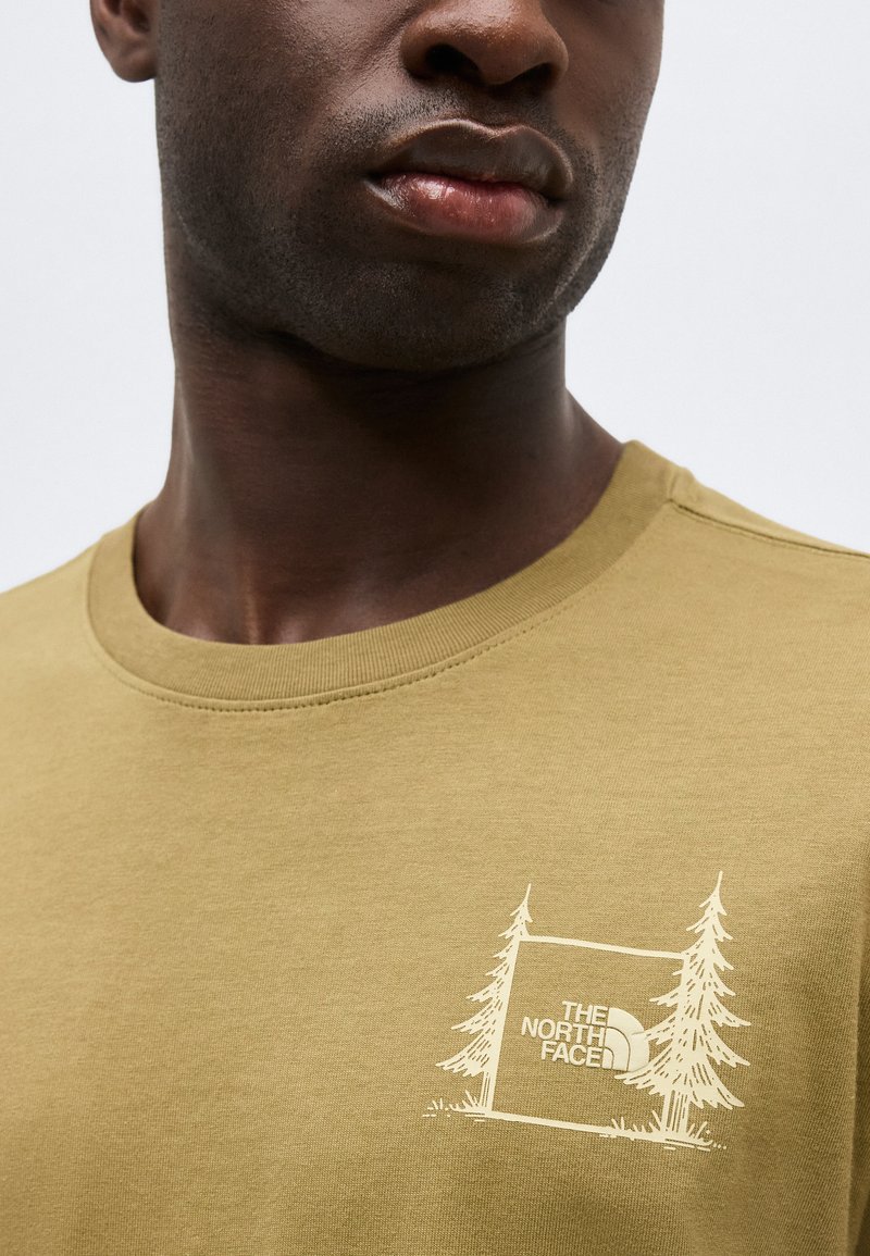 Nahaufnahme einer Person, die ein senfgelbes T-Shirt mit dem weißen Logo "The North Face" und einem Baumgrafik auf der Brust trägt.