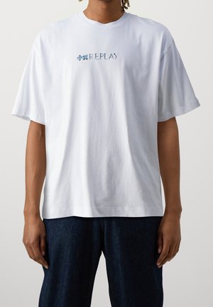 Personne portant un T-shirt blanc ample avec le texte bleu "REPLAY" et des symboles sur la poitrine, associé à un jean bleu foncé, sur un fond uni.