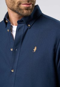 Tamsiai mėlyna mygtukų marškiniai, pagaminti iš tekstūruoto audinio, su auksiniu siuvinėtu logotipu ir kontrastingais mygtukais. Klasikinio apykaklės dizainas.