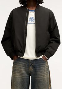 Zwarte bomberjack met geribbelde manchetten, ritssluiting en zijzakken, gedragen over een wit t-shirt en versleten blauwe jeans.