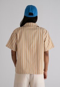 Short-sleeve, collared shirt in light beige with vertical orange stripes, made of lightweight fabric. Worn with a blue cap.